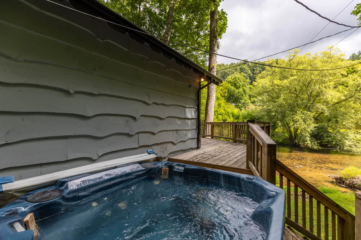 A serene deck featuring a private hot tub framed by leafy trees, with the gentle river flowing nearby. The scene radiates tranquility and intimacy, ideal for unwinding together while feeling secluded in nature.