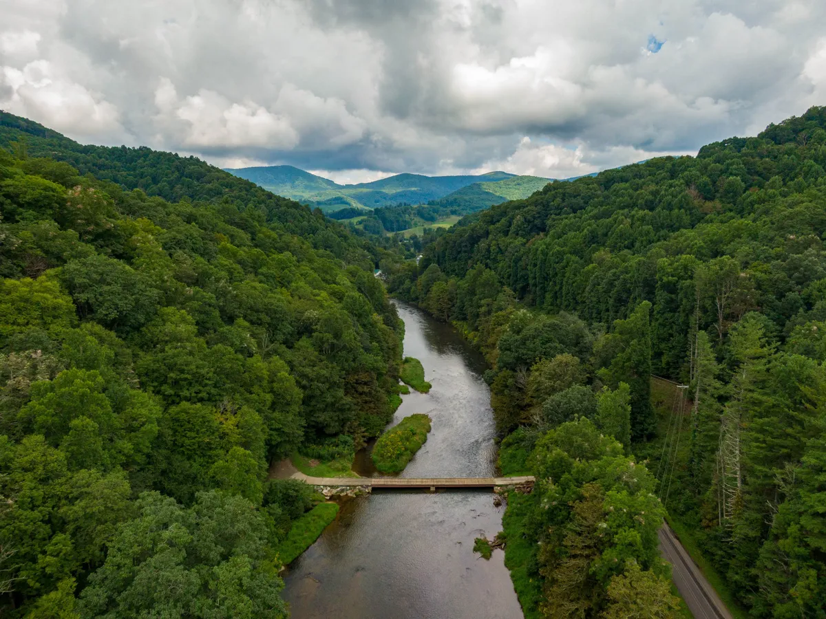 A sweeping view of the river winding through the landscape, bordered by lush green trees and rolling hills. A picturesque bridge spans the water, with open skies and hints of wildlife in the scene, capturing the serenity and adventure potential of the surrounding area.