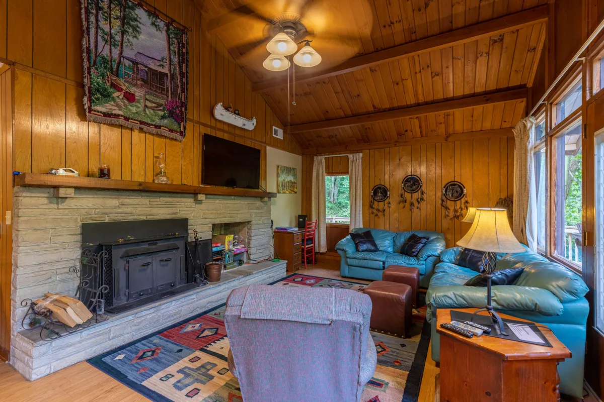 A welcoming living room featuring a glowing fireplace, a blue leather sofa, a patterned rug, and a ceiling fan, all set against warm wood accents. Large windows offer glimpses of the mountain landscape, and the space feels cozy and inviting.