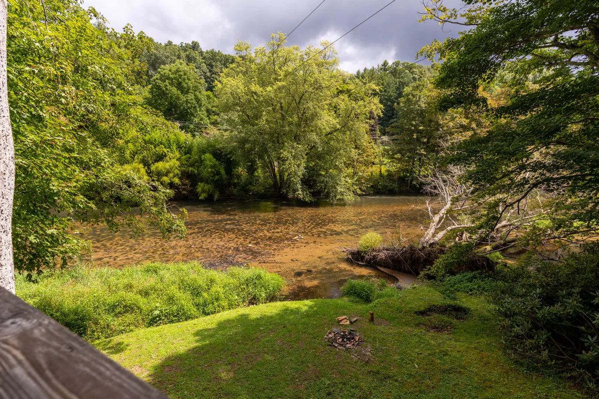A quiet riverfront lawn bordered by trees, with a fire pit in the foreground and the river shimmering in the distance. The area invites peaceful strolls, riverside contemplation, and cozy evenings by the fire.
