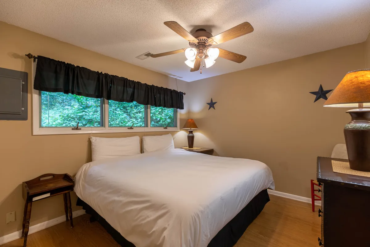 A sunlit bedroom with a comfortable bed, contemporary lamp, large windows, and a ceiling fan. The space exudes calm and features modern accents, with natural light streaming in, ideal for restful sleep or a peaceful afternoon read.