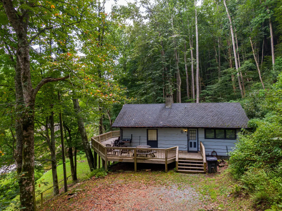 An exterior view of the cozy wooden cabin nestled among trees, with a spacious deck offering outdoor dining and relaxation space. The fire pit is visible in the foreground, and the warm glow of sunset bathes the scene, suggesting evenings spent gathered outdoors.