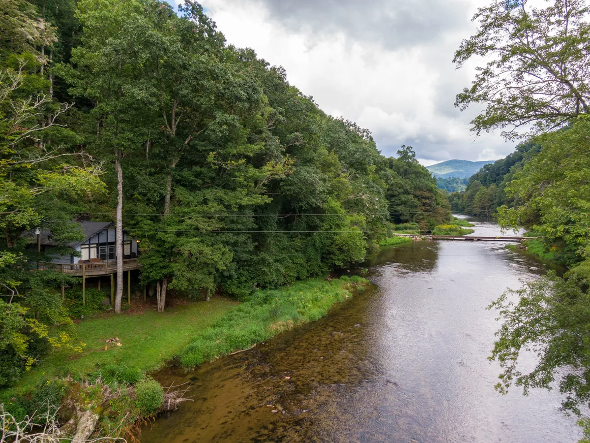 The hero image showcases a charming riverside cabin nestled among towering trees, with the tranquil South Fork of the New River flowing by just steps from the porch. The serene setting is framed by lush greenery, and the cabin's inviting front porch overlooks the water, perfect for relaxing and soaking in the peaceful mountain views.