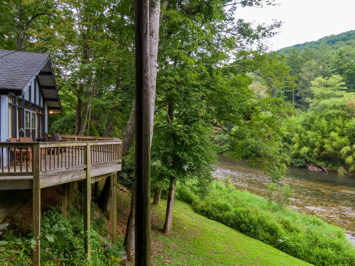 A spacious wooden deck overlooks the flowing river, framed by surrounding trees. The deck is clearly designed for relaxation and gatherings, while the proximity to the water suggests easy access for fishing, floating, or simply enjoying the natural beauty. The mood is tranquil, inviting viewers to imagine themselves sharing laughs or s'mores at sunset.