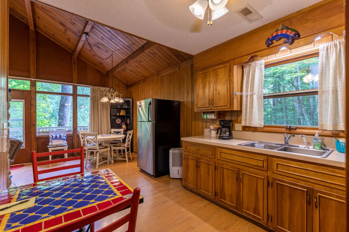 A bright, welcoming dining area with a red and blue patterned table, white chairs, and wooden cabinetry. A large window frames view of the surrounding trees, filling the space with natural light and connecting the indoors to the mountain environment.