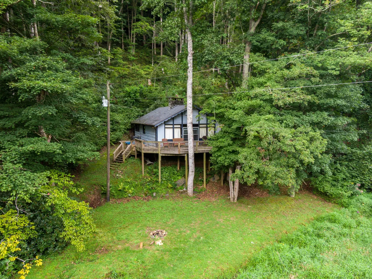 This exterior view captures the house with its expansive deck area, surrounded by mature trees. The fire pit in the yard is clearly visible, and the tranquil scene invites guests to relax on the deck, enjoy the campfire, or take in the natural surroundings. The image showcases the versatility of the outdoor space for morning coffee, stargazing, and social gatherings.