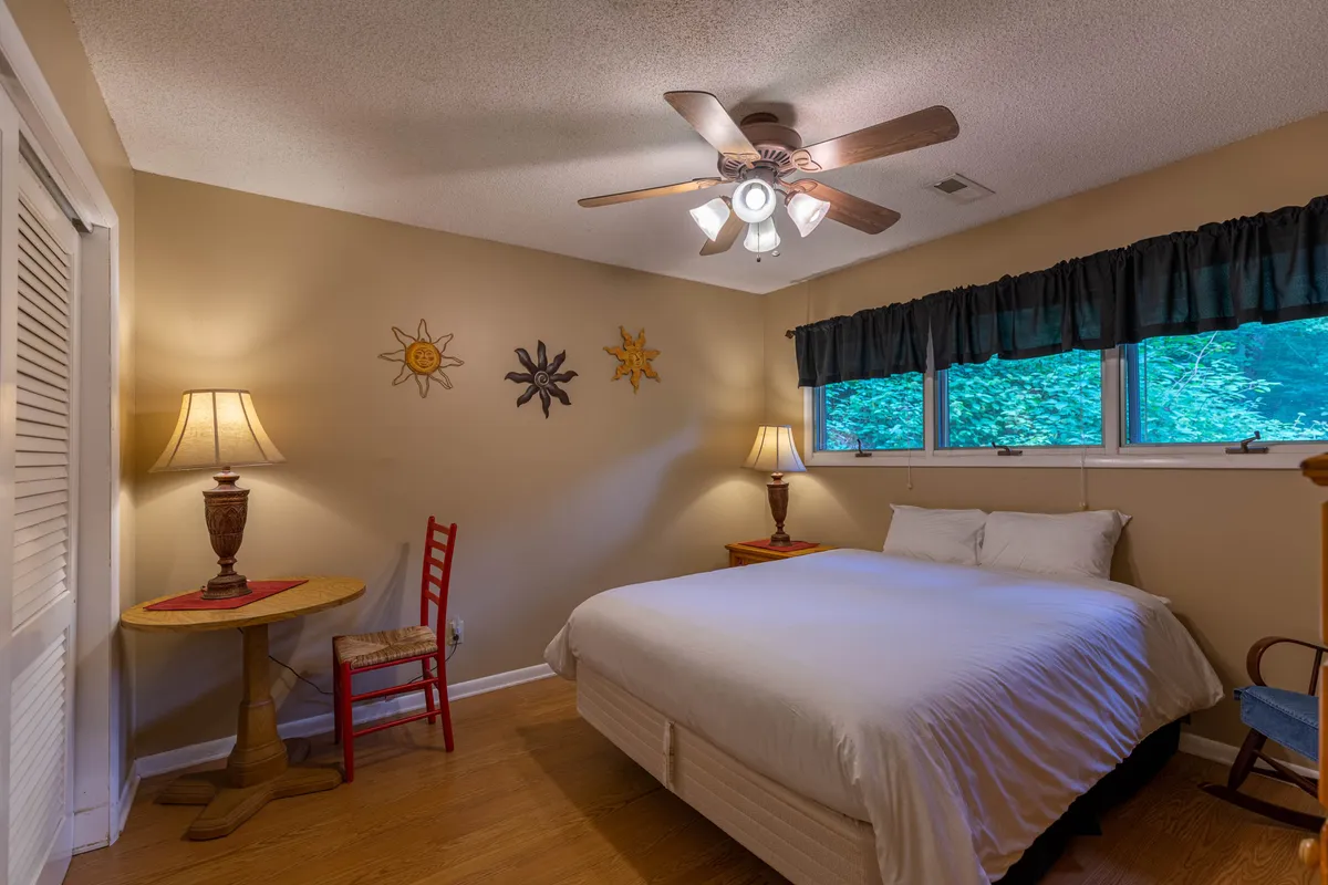 A tranquil bedroom featuring a plush bed, two modern table lamps, a large window, attractive wall art, and a ceiling fan. Soft lighting and stylish décor create a soothing environment ideal for relaxation and rejuvenation.