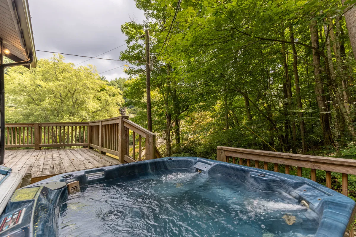 This image features a private hot tub set on a wooden deck, surrounded by lush trees, offering a tranquil and secluded ambiance. The placement of the hot tub ensures privacy, while the deck provides additional space for relaxation and entertaining guests. The peaceful mood is enhanced by the natural backdrop, which hints at the proximity to the river and the calming presence of nature.