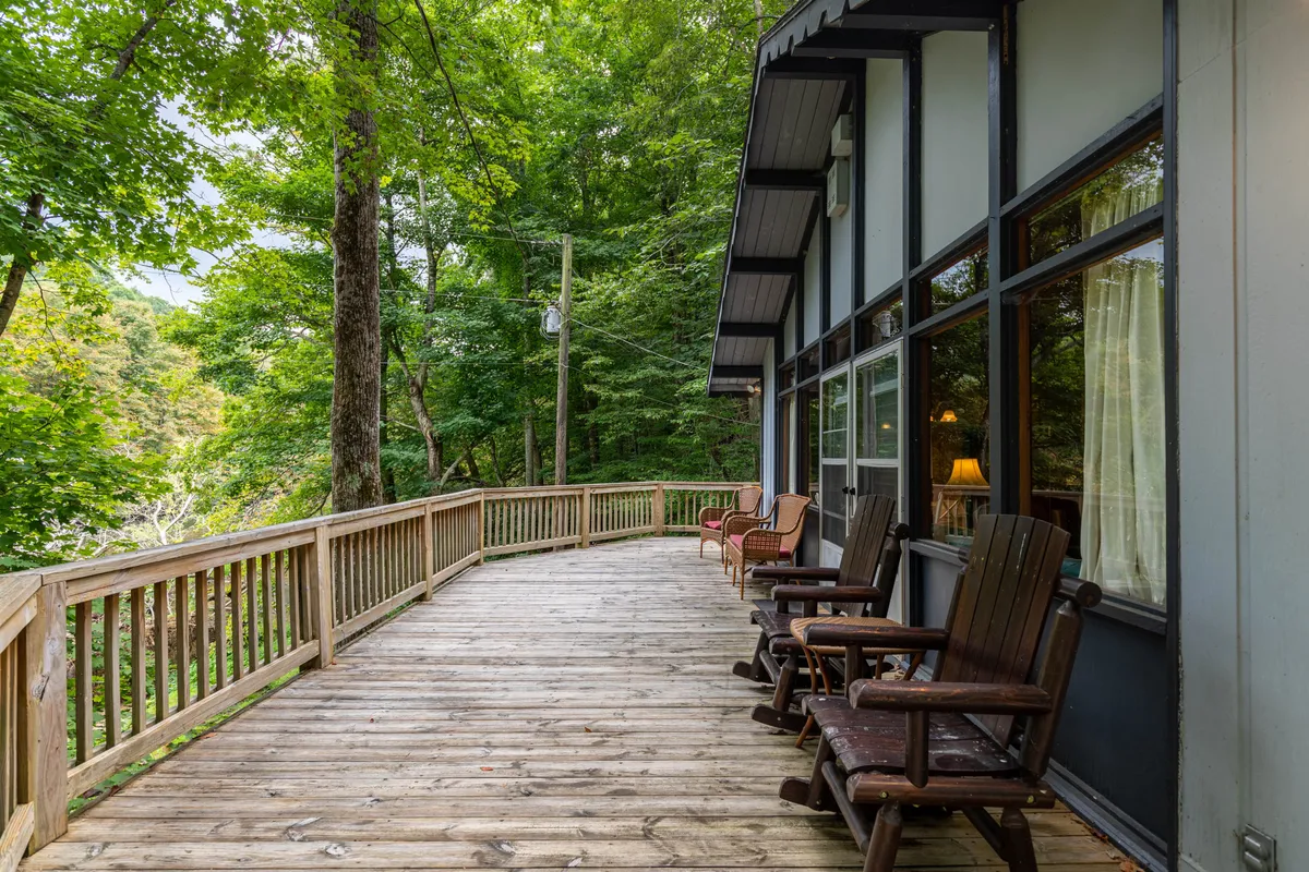 A peaceful wooden deck shaded by trees, with classic rocking chairs positioned to overlook the tranquil natural surroundings. The setting suggests a perfect spot for sipping coffee in the morning or sharing a quiet moment while immersed in nature.