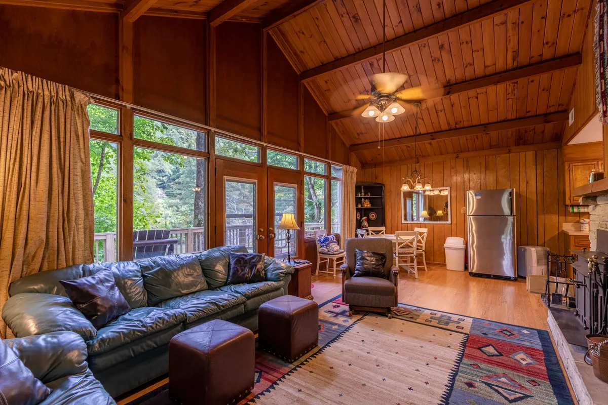 A combined living and dining space with a leather sofa, wooden ceiling, large windows overlooking the woods, a rug, and a visible dining area. The arrangement is ideal for relaxing, dining, and enjoying the tranquil natural surroundings from indoors.