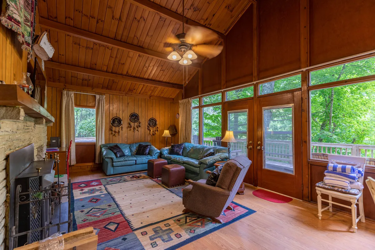A cozy great room with a prominent stone fireplace, comfortable seating arranged around it, a colorful area rug, and expansive windows that offer views of the surrounding woodland. The modern furnishings invite relaxation and gathering, while the natural light brings the outdoors in.