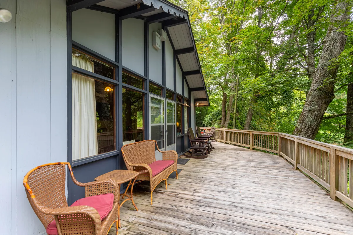 A tranquil exterior shot featuring a wooden deck with wicker chairs, surrounded by greenery. The space is set up for relaxation and socializing, with the natural landscape providing a peaceful backdrop, perfect for morning coffee or evening conversation.
