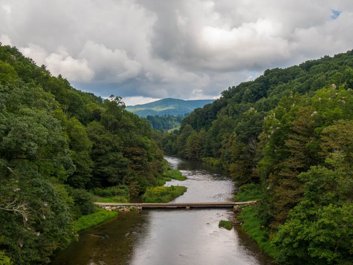 A scenic view of the river, bridge, and distant mountains framed by trees. The bridge and river are focal points, creating a strong sense of place and security, with natural beauty inviting exploration.